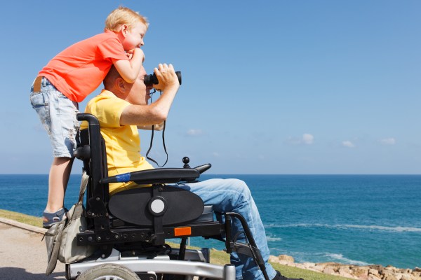 Disabled father, sat in powered wheelchair looking out to sea with a pair of binoculars with a young boy with blonde hair standing on the back of his wheelchair smiling